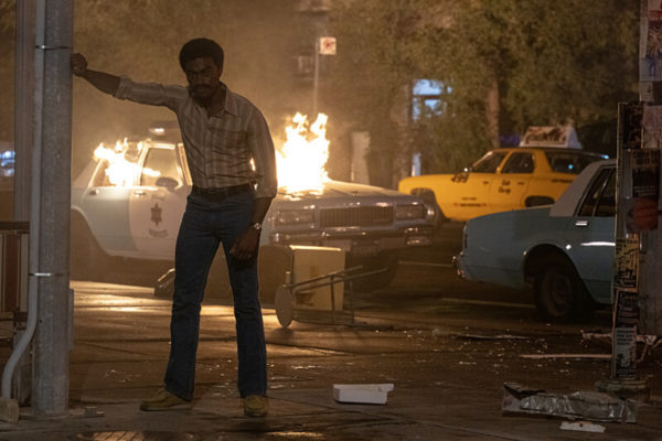 A black man leans against a wall in front of a police cruiser on fire